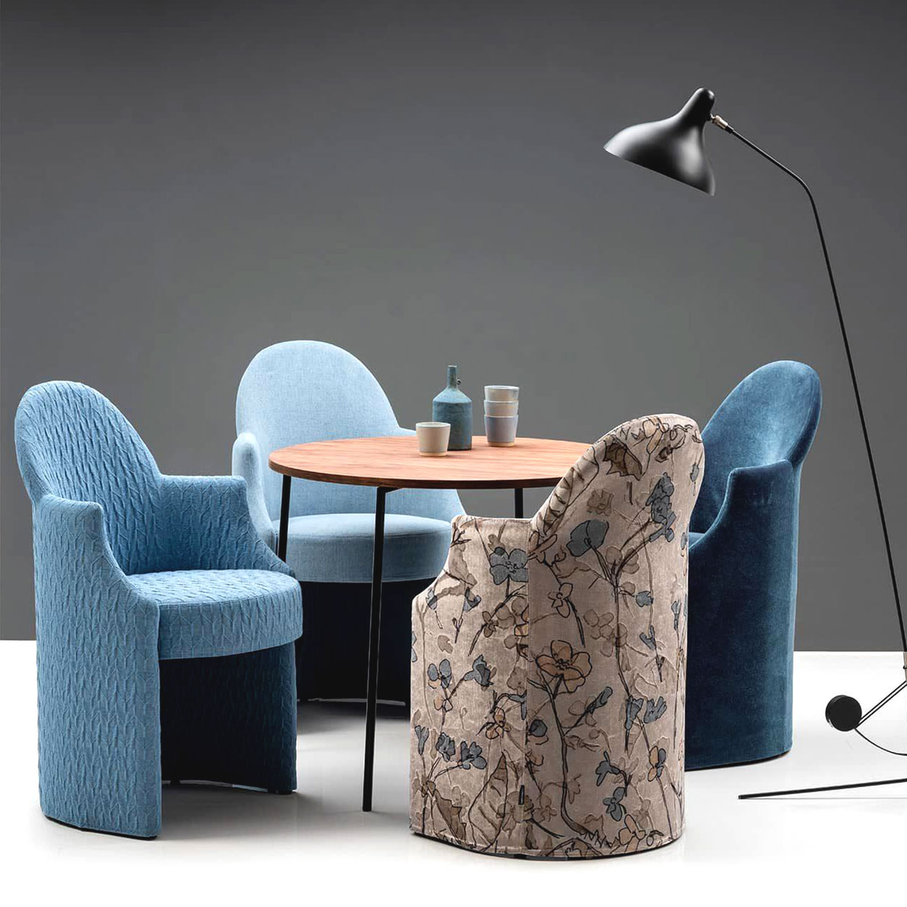 brühl armand Sessel 71601 Vier Sessel stehen um einen runden Tisch mit Holzplatte und schwarzem Metalluntergestell. Drei der Sessel sind in verschiedenen Blautönen bezogen, der Sessel vorne rechts ist mit einem floralen Muster in hellbraun und blau bezogen.