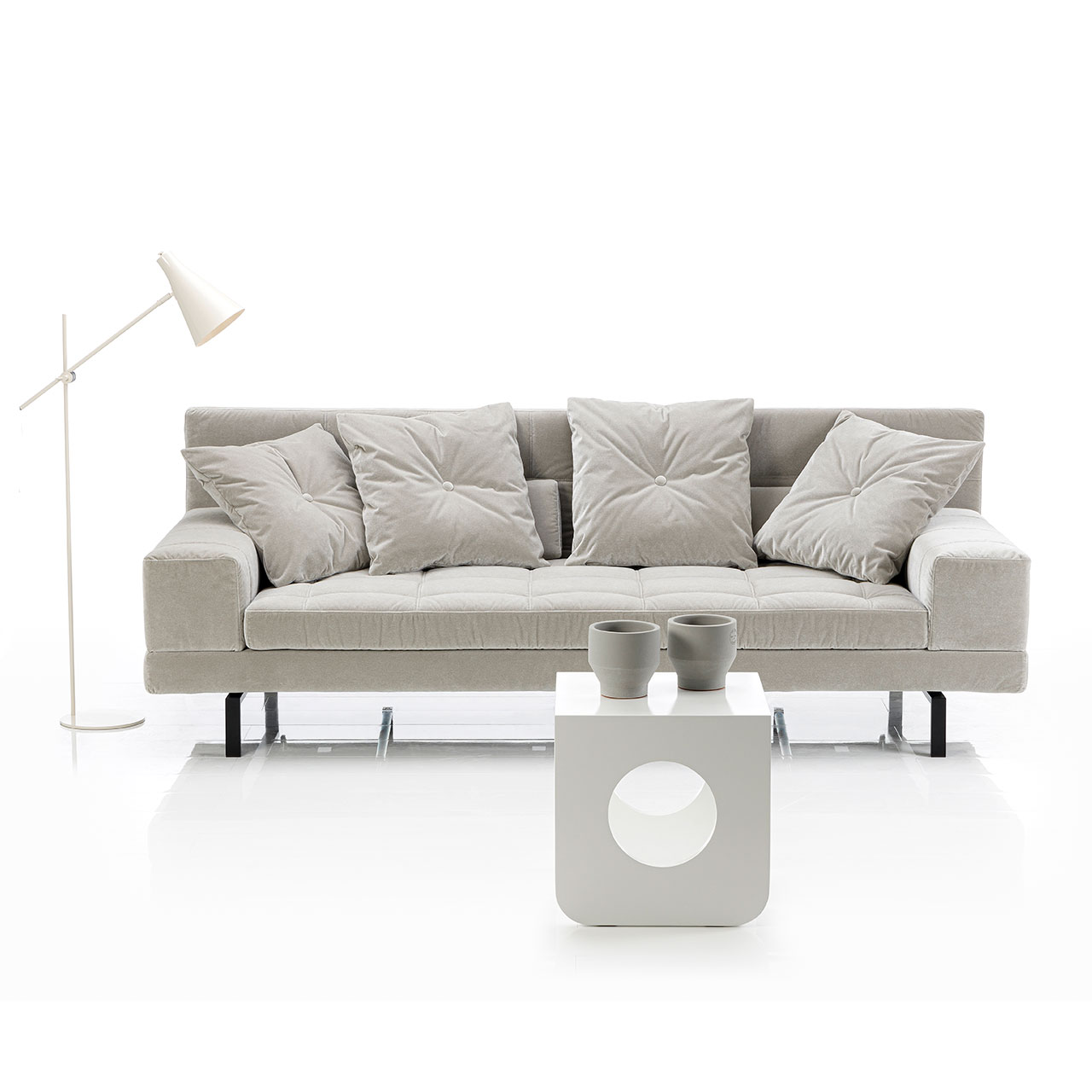 brühl amber Sofa 3-Sitzer Ambientebild: Graues Sofa mit niedrigen, breiten Armlehnen. Auf dem Sofa liegen vier quadratische Kissen. Vor dem Sofa steht ein weißer Beistelltisch.