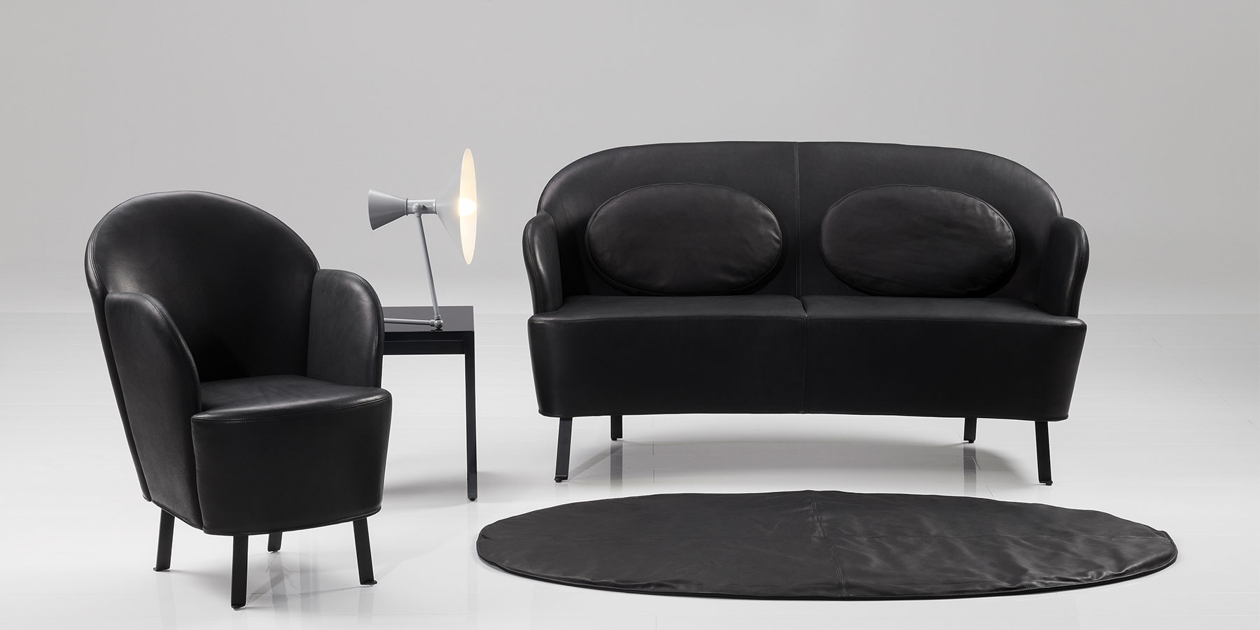 Sofas und Sessel von brühl floret Ein schwarzes Ledersofa mit gerundeter Rückenlehne und filigranen schwarzen Metallfüßen steht neben einem ebenfalls schwarzen Ledersessel mit rundem Rücken.