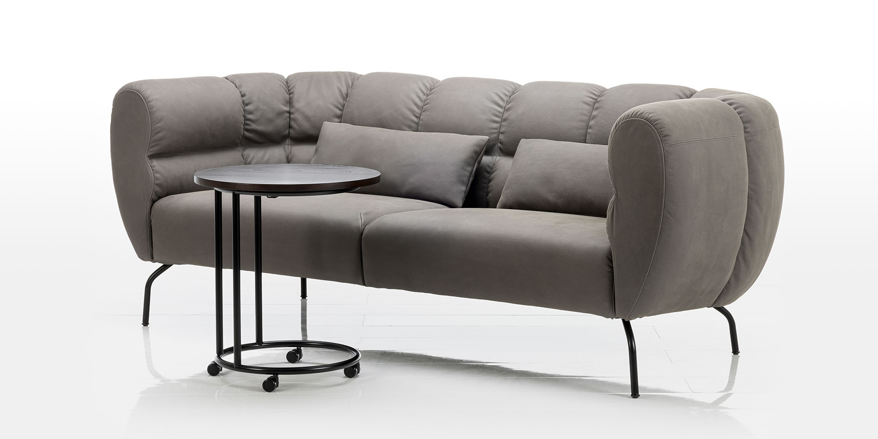 Tische von brühl Graues Sofa der Serie magnolia. Davor steht ein runder Beistelltisch mit kleinen Rollen.