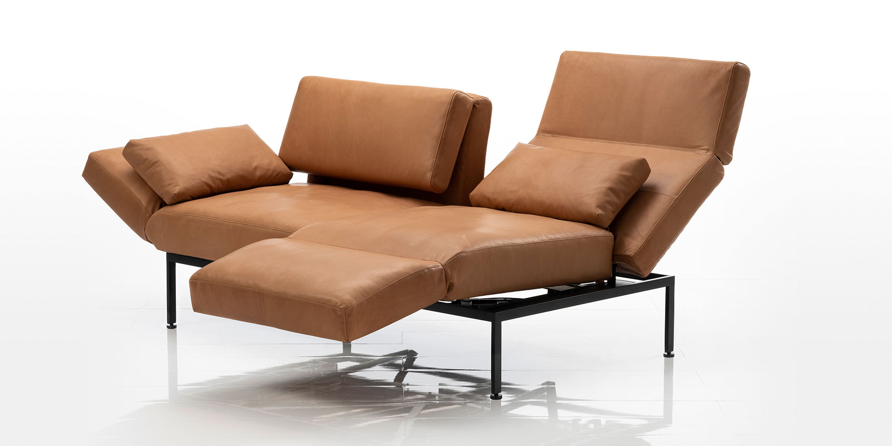 Multifunktionssofas brühl roro soft Ein zweisitziges Sofa in braunem Leder. Am rechten Sitzplatz wurde die Armlehne zur Fußstütze umgeschwenkt, um eine Relaxposition herzustellen. Die linke Armlehne wurde leicht abgesenkt.
